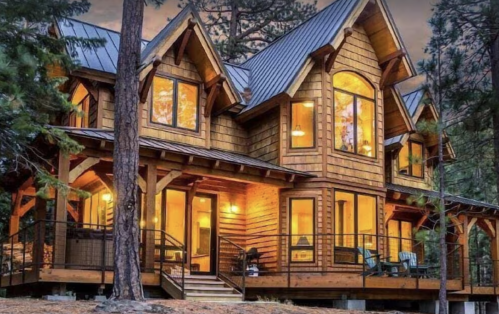 A cozy wooden cabin with large windows, surrounded by trees, illuminated at dusk.