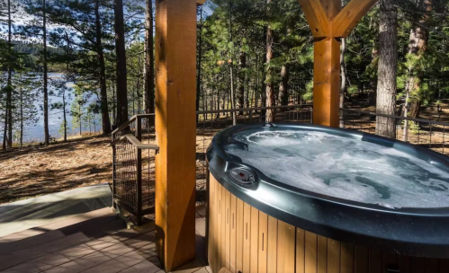 A hot tub on a wooden deck surrounded by trees, overlooking a serene lake in a natural setting.