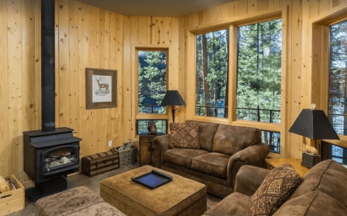 Cozy living room with wooden walls, large windows, a wood stove, and comfortable brown furniture.