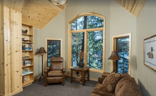 Cozy living space with wooden accents, large windows, and a view of trees, featuring a sofa and bookshelves.