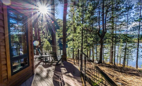 A sunlit deck surrounded by tall trees, overlooking a serene lake in a forested area.