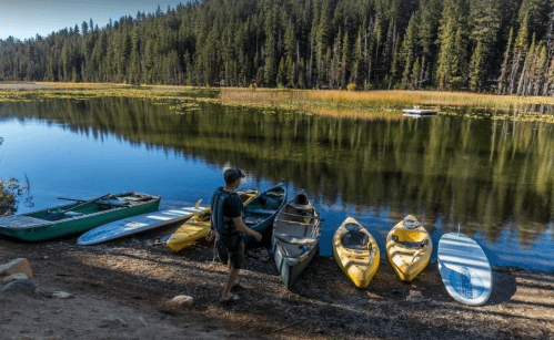 A person stands by a calm lake with several kayaks and a paddleboard lined up on the shore, surrounded by trees.