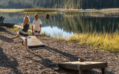 Two children play cornhole by a serene lake, surrounded by trees and autumn foliage.