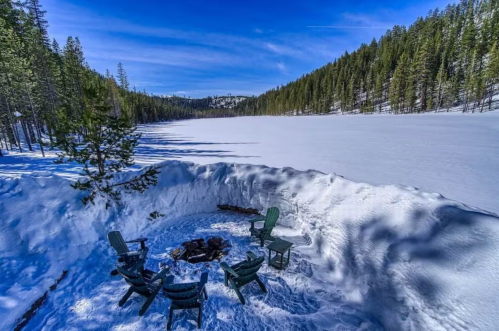 A snowy landscape with a frozen lake and a fire pit surrounded by green chairs, framed by tall pine trees.