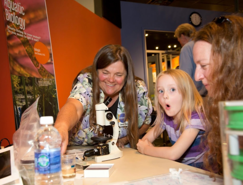 A woman shows a child a microscope while another adult watches, all engaged in a science activity.