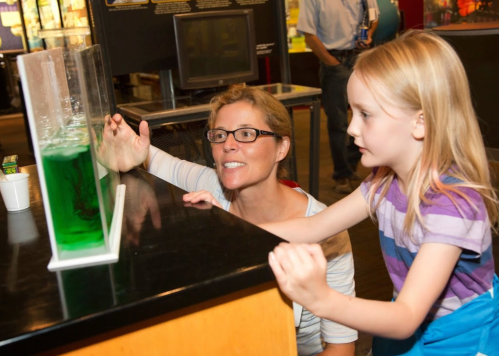 A woman and a girl observe a green liquid in a display at a science museum, both engaged and curious.
