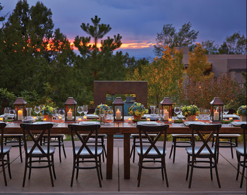 A beautifully set outdoor dining table at sunset, surrounded by greenery and lanterns, ready for a gathering.