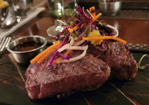 A plate of grilled steak topped with colorful shredded vegetables and a small bowl of sauce on the side.
