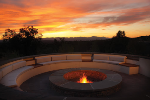 A circular outdoor seating area with a fire pit, set against a vibrant sunset and distant mountains.
