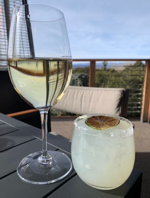 A glass of white wine and a cocktail with a lime slice on a table, with a scenic outdoor view in the background.