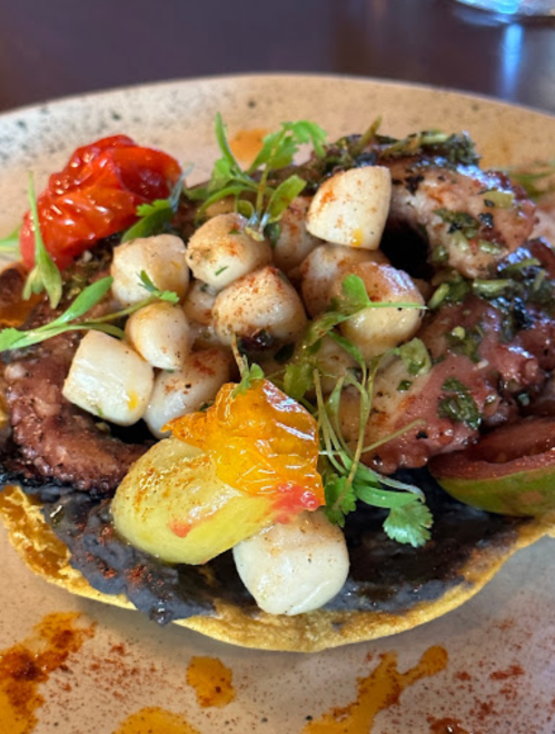 A colorful plate featuring a tostada topped with grilled octopus, scallops, cherry tomatoes, and fresh herbs.