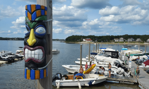 A colorful tiki mask on a post overlooks a busy marina with boats and people enjoying the water on a sunny day.