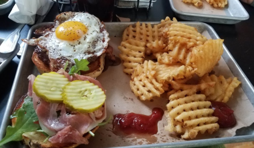A tray with a burger topped with a fried egg, lettuce, ham, pickles, and waffle fries with ketchup on the side.