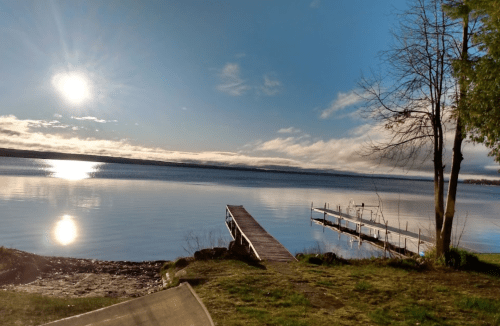 A serene lake scene with a wooden dock, reflecting sunlight and surrounded by trees under a clear blue sky.
