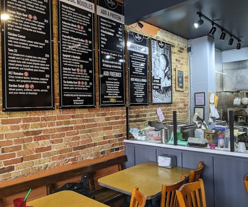 Interior of a casual restaurant featuring a brick wall, menu boards, and wooden tables.