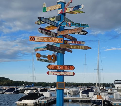 A colorful signpost with directional arrows pointing to various cities and attractions, near a marina with boats.