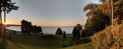 A serene sunset over a calm lake, framed by trees and lush greenery in the foreground.