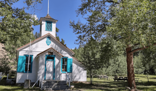 A quaint white church with teal shutters, surrounded by trees and picnic tables in a serene outdoor setting.
