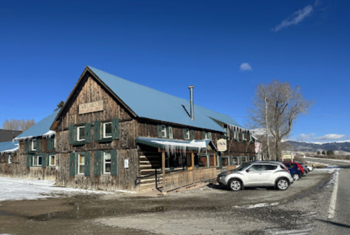 A rustic wooden inn with a blue roof, surrounded by snow, and parked cars in front under a clear blue sky.