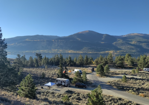 A scenic view of a campsite by a lake, surrounded by mountains and trees under a clear blue sky.