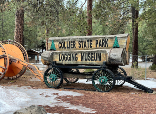 A wooden wagon with logs displays a sign for Collier State Park Logging Museum, surrounded by trees.