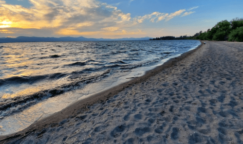A sandy beach by a lake at sunset, with gentle waves and distant mountains under a colorful sky.
