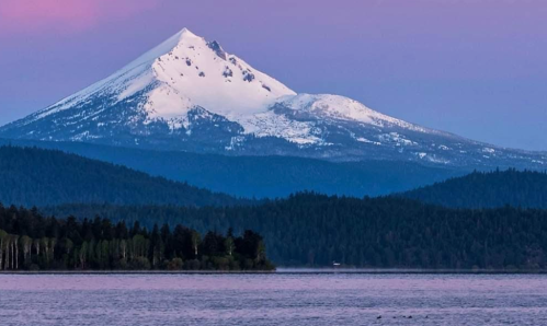 Snow-capped mountain rises above a serene lake, surrounded by lush forests under a pastel sky.