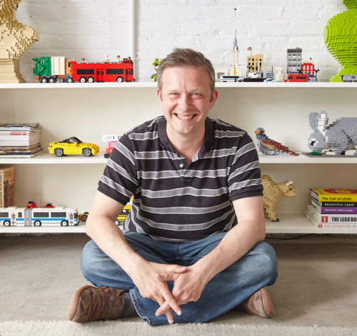 A smiling man sits cross-legged on the floor in front of a shelf filled with colorful LEGO creations and books.