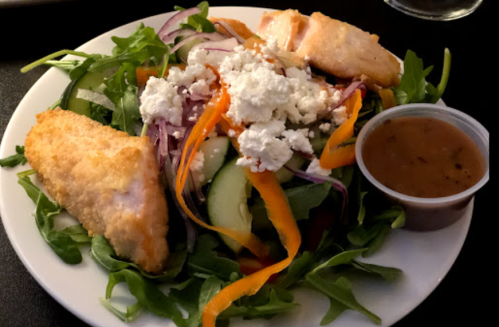 A plate of salad with mixed greens, grilled salmon, sliced vegetables, feta cheese, and a side of dressing.