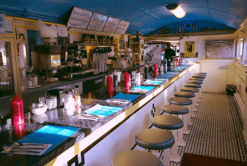 A retro diner interior featuring a long counter with stools, condiments, and a colorful menu on the wall.