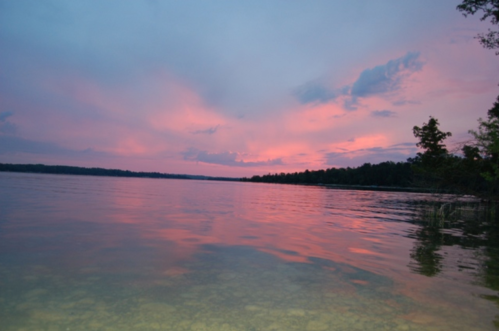 A serene lake at sunset, reflecting pink and purple hues in the water, surrounded by trees and a calm sky.