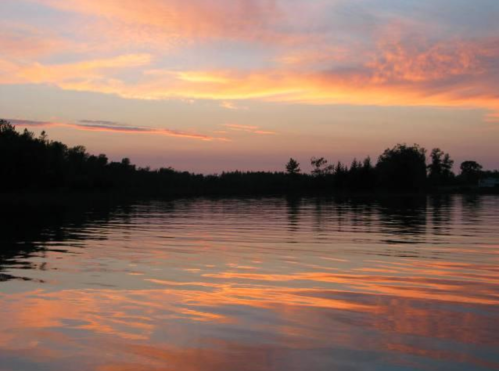 A serene lake at sunset, reflecting vibrant orange and pink hues in the water, surrounded by trees.