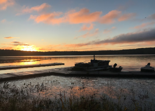 Sunset over a calm lake with boats docked and reflections on the water, surrounded by trees and a colorful sky.