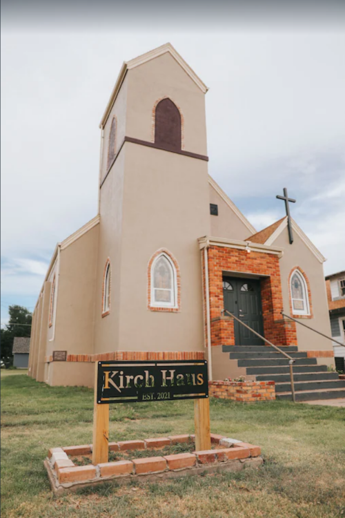 A beige church building with a cross, featuring arched windows and a sign reading "Kirch Haus, EST. 2021."