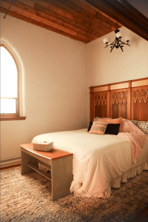 Cozy bedroom with a wooden bed, decorative pillows, and a soft rug under a sloped ceiling with an arched window.