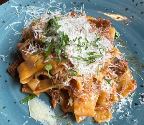 A plate of pappardelle pasta topped with a rich sauce, grated cheese, and fresh herbs on a blue dish.