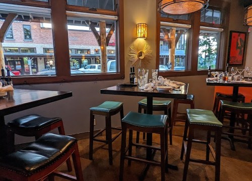 Cozy restaurant interior with wooden tables, high stools, and large windows overlooking a street.