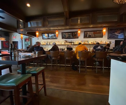 A cozy bar scene with patrons seated at the counter, shelves lined with bottles, and warm lighting.