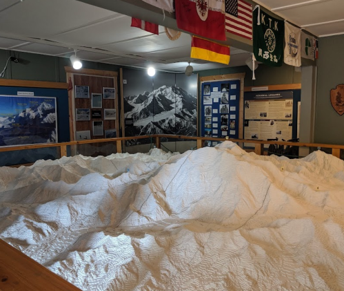 A detailed topographical model of mountains displayed in a museum, with informational panels and flags in the background.