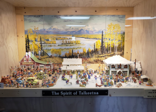 A detailed display of miniature figures and buildings depicting a winter scene in Talkeetna, Alaska, with a scenic backdrop.