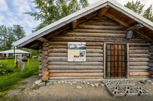 Log cabin exterior with a wooden door, a sign on the wall, and a grassy area in the background.