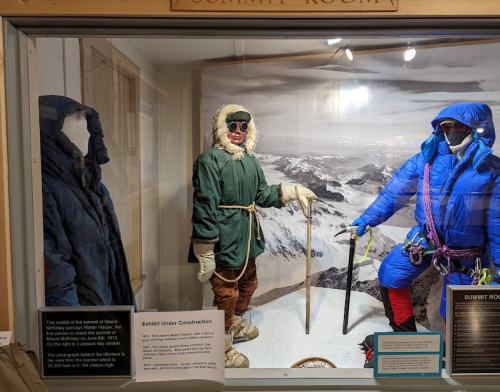 Display of mountaineering gear in a glass case, featuring two figures in winter clothing on a snowy mountain backdrop.