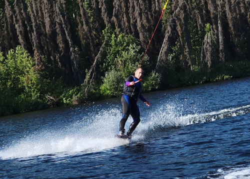A person water skiing on a lake, creating splashes, with lush greenery in the background.