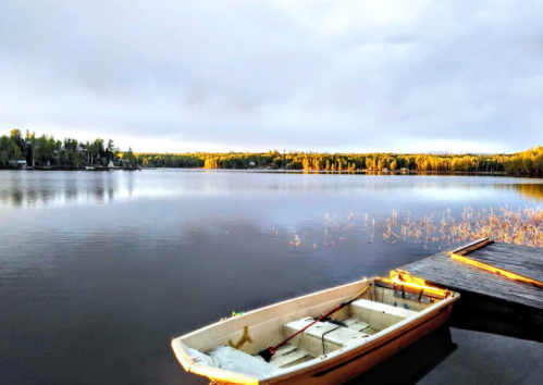 A calm lake at sunset with a small boat docked and trees lining the shore under a cloudy sky.