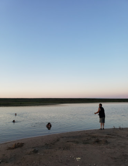 A person stands on the shore of a calm lake at sunset, while another person swims nearby.