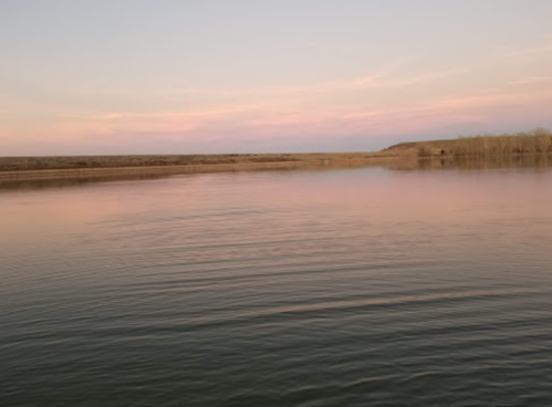 A calm river at sunset, reflecting soft pink and purple hues in the sky, with trees lining the distant shore.