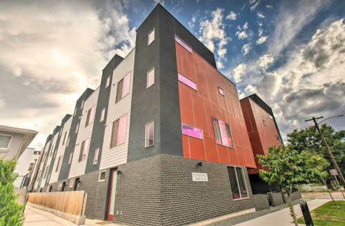 Modern multi-story building with a mix of dark and light panels, large windows, and a cloudy sky in the background.