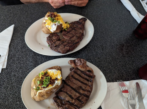 Two plates of grilled steak with baked potatoes topped with sour cream, cheese, and green onions on a table.