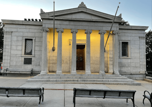 A stone building with columns and a yellow-lit entrance, flanked by benches and surrounded by trees.