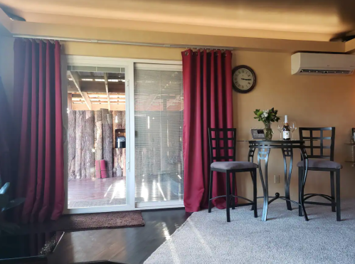 Cozy interior with a small dining table, two chairs, red curtains, and a view of a wooden deck through sliding doors.